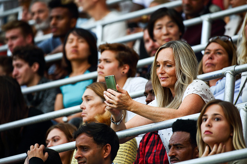 Full arena of people waiting for the sport match to start. Large group of people, audience at the stadium. Caucasian woman looking at her phone.