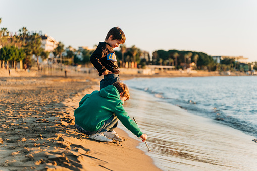 Two school boys kids playing having fun with winter sea waves on the sea shore. Schoolchildren friends playing with sand near autumn ocean waves during a mild fall sunset.