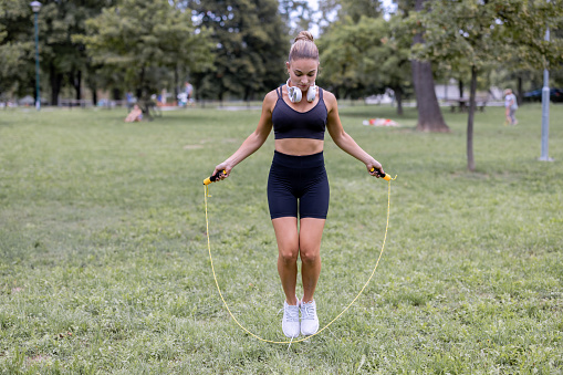 Beautiful focused Caucasian athlete skipping jumping rope in nature with white headphones around her neck