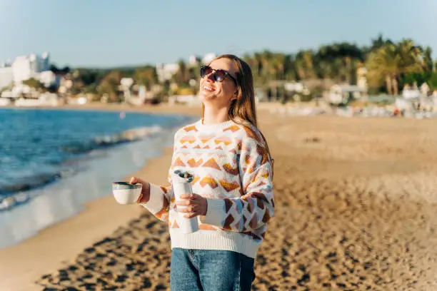 Young beautiful woman in cozy sweater and sunglasses enjoying hot coffee from thermo mug while relaxing on winter seaside sand beach. Cute attractive girl drinks tea taking sunbathe near ocean.