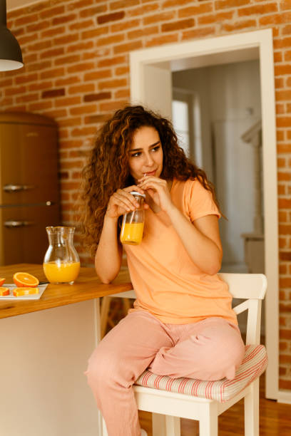 joven y hermosa mujer sentada en la cocina y bebiendo jugo de naranja recién exprimido - drinking straw drinking juice women fotografías e imágenes de stock