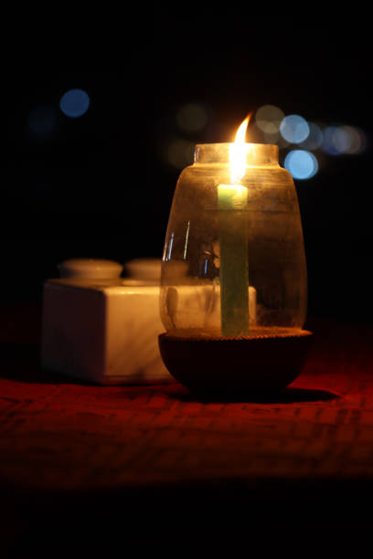 Stock photo showing close-up, night-time view of burning candle flame in a glass and coconut shell candle holder on tablecloth covered al fresco dining table on sandy beach.