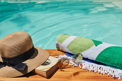 Closeup, sunglasses and book by swimming pool for relax holiday, vacation or summer break. Luxury, towel and fashion accessories by outdoor water space for getaway, free time and travel resort