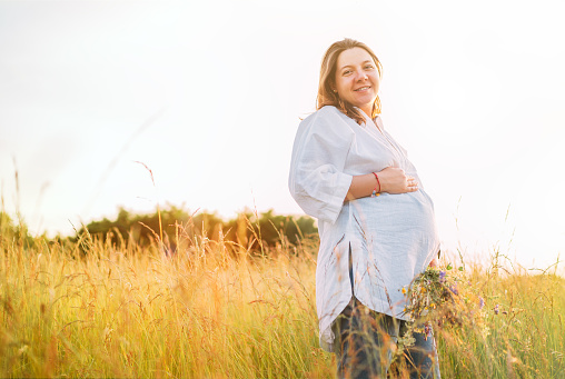 Sincerely smiling young pregnant woman dressed in light summer clothes walking by the high green grass meadow with wildflowers bouquet in evening sunset hours. Human in the nature concept image.