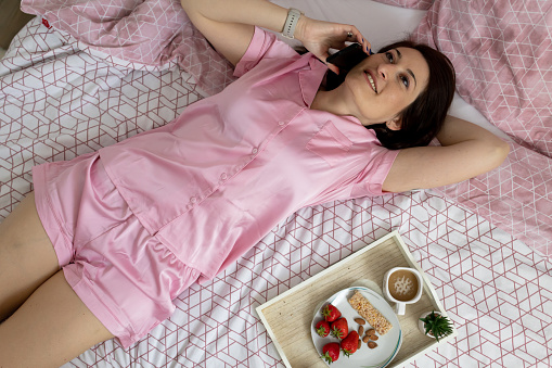 Woman enjoying warm coffee and talking on telephone in her bedroom at home. She has cereal bar and strawberry for breakfast. Fruit and nut, muesli bar.