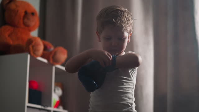 Focused toddler puts on boxer glove fixing on hand in room