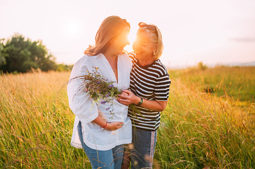 Portrait of a sincerely smiling young pregnant woman dressed in light summer clothes embracing with mother in evening sunset hours. Woman's health, human in the nature concept image.