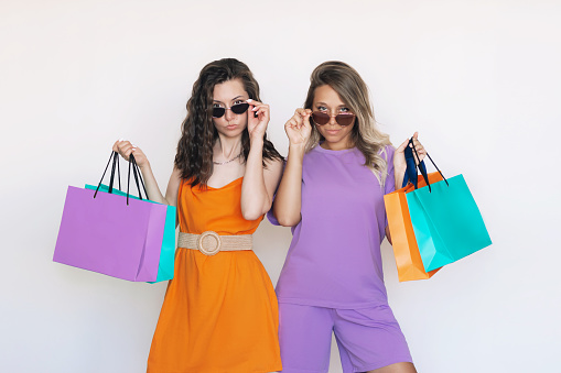 Two young attractive caucasian blonde and brunette women in bright colored clothes and sunglasses hold paper color eco bags in their hands isolated on a white background. Shopping and fashion concept