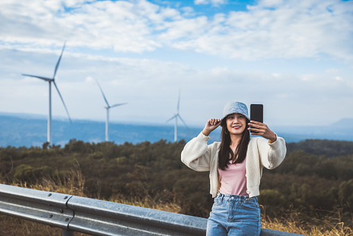 Travel on bike concept, Happy asian woman traveler relax and selfie from mobile phone with group of Wind turbine on mountain background in Lam Takong Dam, Nakhon Ratchasima, Thailand