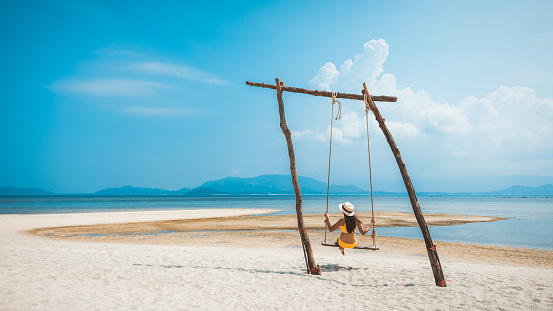 Summer travel vacation concept, Happy traveler asian woman with bikini and hat relax in swing on beach in Koh Rap, Samui, Surat Thani, Thailand