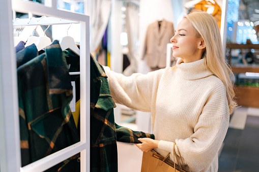 Side view of pretty young woman choosing coat in clothing store, standing near showcase with female clothing, sorting through different options on hanger. Concept of holiday sales, retail.