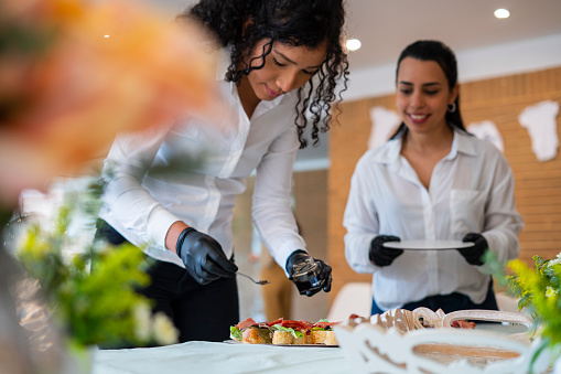 Average age 30-year-old Latina women dressed in white shirts and pants, professional waitresses are inside the facilities of a Veneto organizing the decoration and food table