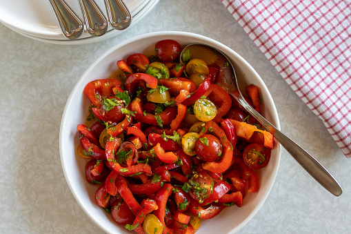 Delicious and healthy homemade summer salad with red and yellow cherry tomatoes and bell peppers marinated with honey, lemon juice, olive oil and chopped parley. Served on white background from above in a bowl with spoon and napkin