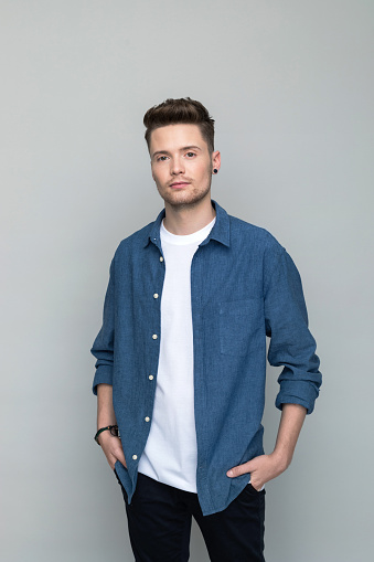 Handsome young man wearing denim shirt and white t-shirt standing with hands in pockets and looking at camera. Studio shot, grey background.