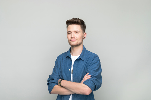 Handsome young man wearing denim shirt and white t-shirt standing with arms crossed and looking at camera. Studio shot, grey background.