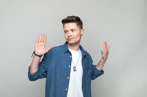 Handsome young man wearing denim shirt and white t-shirt standing with raised hands and looking away. Studio shot, grey background.