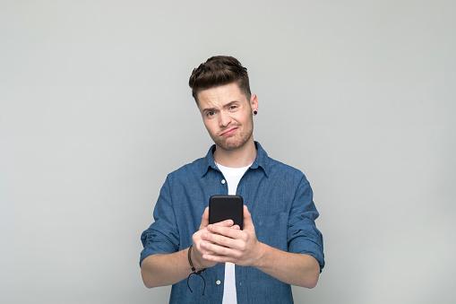 Disappointed young man wearing denim shirt and white t-shirt holding mobile phone. Studio shot, grey background.