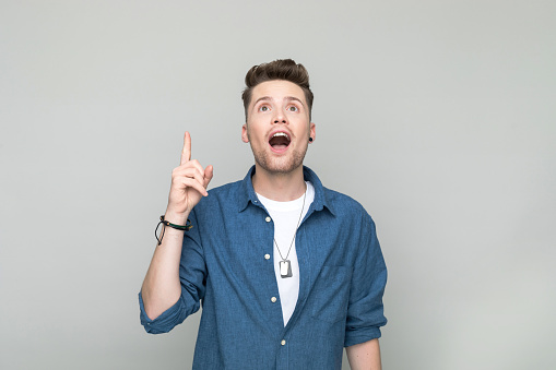 Surprised young man wearing denim shirt and white t-shirt pointing with index finger at copy space and looking up. Studio shot, grey background.