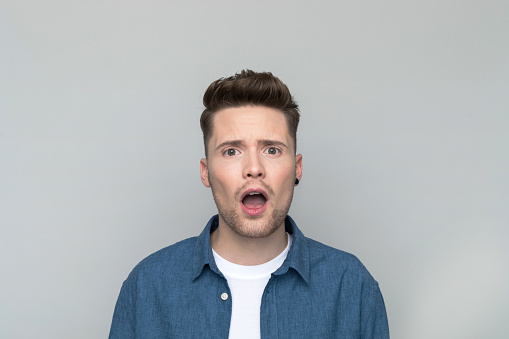 Headshot of shocked young man wearing denim shirt and white t-shirt staring at camera with mouth open. Studio shot, grey background.