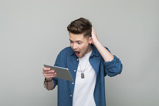 Excited young man wearing denim shirt and white t-shirt holding iPad with hand in hair. Studio shot, grey background.