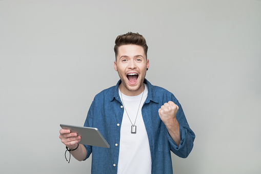 Excited young man wearing denim shirt and white t-shirt holding iPad. Studio shot, grey background.
