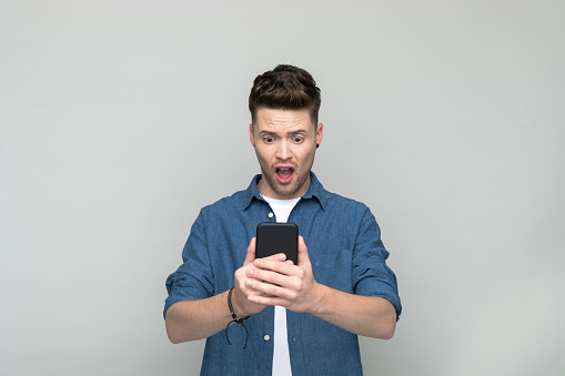 Shocked young man wearing denim shirt and white t-shirt holding mobile phone in both hands. Studio shot, grey background.