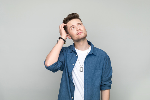 Worried young man wearing denim shirt and white t-shirt looking up with hand in hair. Studio shot, grey background.