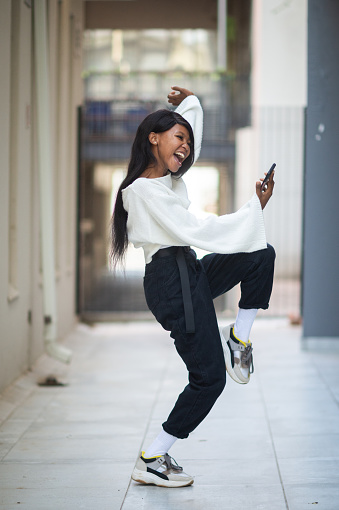 Jumping arm raised Young African woman with long black hair showing surprise and happiness getting good news on her smart phone.