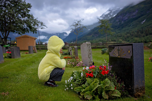 Sad little child, blond boy, standing in the rain on cemetery, sad person, mourning