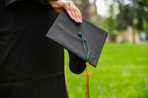 A hand of a woman in a graduation gown holding a morarboard in a park after graduation.