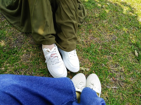 Two pairs of female legs wearing white shoes sneakers with green skirt and denim jean on the grass