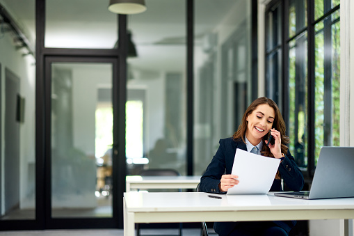 A cheerful pretty female employee holding documents and sitting in front of the laptop while talking on a mobile phone.