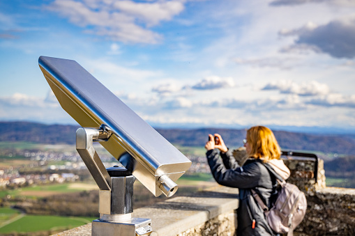 A young woman is on a tourist observation deck with a modern telescope of the medieval castle of Hochnetwil, and takes pictures on her phone of the industrial city of Singen and the cloudy sky
