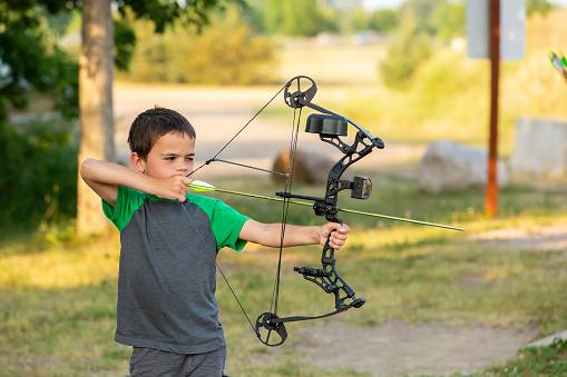 Six year old boy practicing archery at an outdoor range on a summer evening.