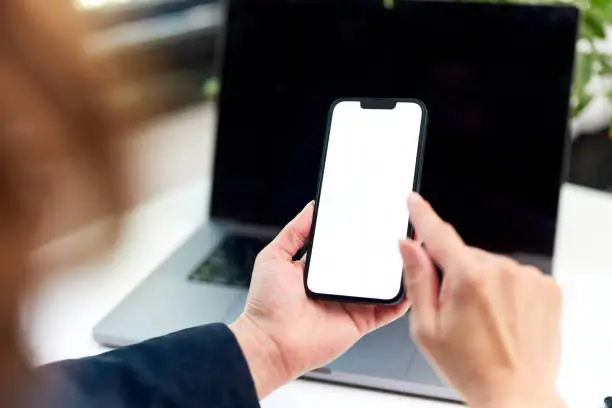 A woman holding a mobile phone with an empty white screen while sitting in front of the turned-off laptop.