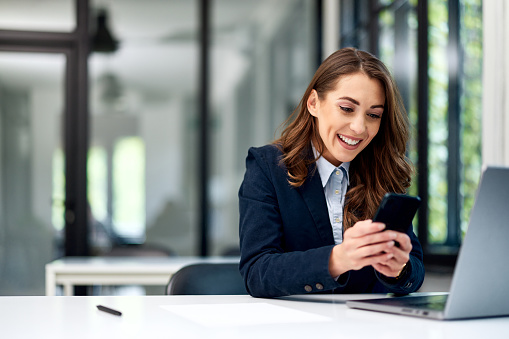 A pretty brunette female employee using a mobile phone and smiling while sitting in front of a laptop at the workplace.