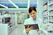 A pretty female pharmacist using a tablet while standing in a drugstore.