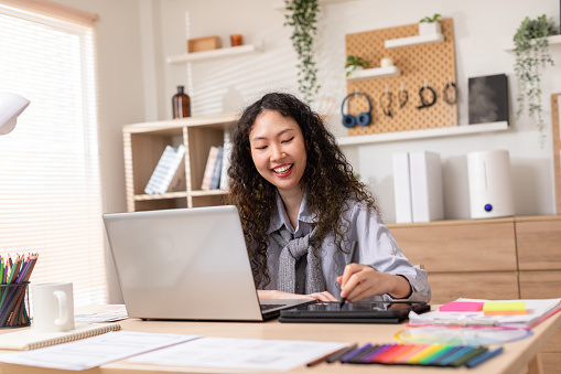 Asian business owner woman smile using stylus pen drawing design her work in home office. Confident designer or creative woman work on laptop. Successful millennial female small business owner concept