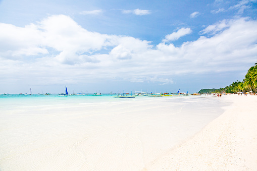 A view of Boracay's White Beach in the Philippines. The image showcases the renowned beach with its distinctive white sand and shallow, transparent waters. Towards the bottom of the frame, the water appears white, revealing the visible sandy ocean floor. Moving towards the center, the water transitions into shades of turquoise and blue along the horizon line. In the distance, several white boats are anchored near the beach. To the right of the frame, a shoreline adorned with palm trees adds a natural element to the scene. The sky above is blue, adorned with scattered clouds. This photograph provides a glimpse of the captivating landscape of Boracay's White Beach, featuring its crystal clear waters, anchored boats, and the presence of palm trees along the shoreline.