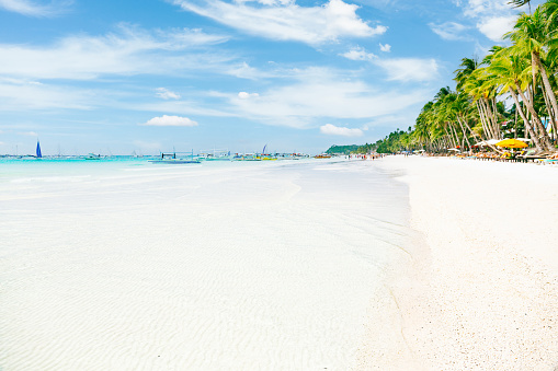 A view of Boracay's White Beach, located in the Philippines. The image showcases the distinct features of the beach, including its pristine white sand and clear, shallow waters. Towards the lower part of the frame, the water appears white, revealing the visibility of the sandy ocean floor. Moving towards the center, the water transitions into turquoise and blue hues along the horizon. In the distance, several boats can be seen anchored near the beach, while a palm-lined shoreline is visible on the right side of the frame. The sky above is adorned with scattered clouds against a blue backdrop. This photograph provides an unadorned depiction of Boracay's White Beach, highlighting its clear waters, anchored boats, and the presence of a shoreline adorned with palm trees.
