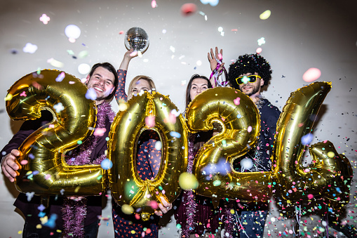 Standing in a row and holding 2024 gold foil balloons, four friends are partying in the New Year amid falling confetti