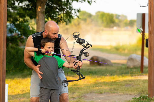 Father teaching his six year old son how to shoot bow and arrow at an outdoor archery range on a summer evening.