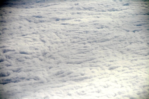 Aerial view of white, fluffy clouds against a light blue sky, seen through an airplane window