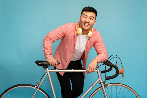 Smiling young man poses leaning against a bicycle in studio, casually dressed against blue background.