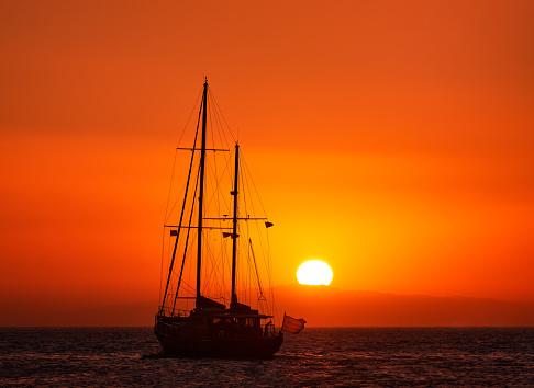 Silhouette of sailing boat with sails down against sun at sunset, sun glare on sea waters. Romantic seascape. Sailboat go to sea, rear view, sun setting in hazy background. Meditative image