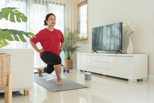 Senior Asian woman exercising and communicating with smart speaker in living room at home. Sport healthy concept.