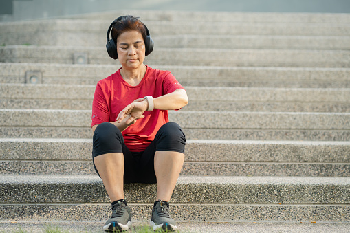 Senior Asian woman wearing headphones exercising and checking smart watch at park. Sport healthy concept.