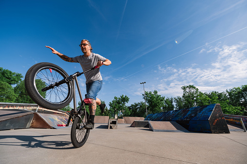 A mature tattooed urban man is performing tricks on a bmx bike while balancing on one wheel in a skate park. A middle-aged extreme man is practicing freestyle tricks on his bike.
