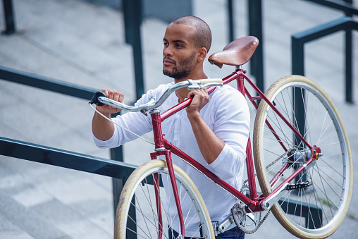 Handsome young businessman in casual clothes is looking away and thinking while holding a bike, standing outdoors