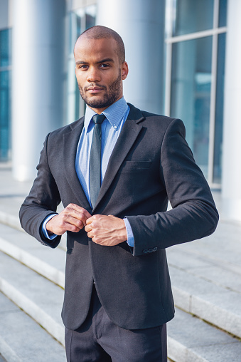 Handsome young businessman in classic suit is buttoning his jacket and looking at camera, standing outside the office building